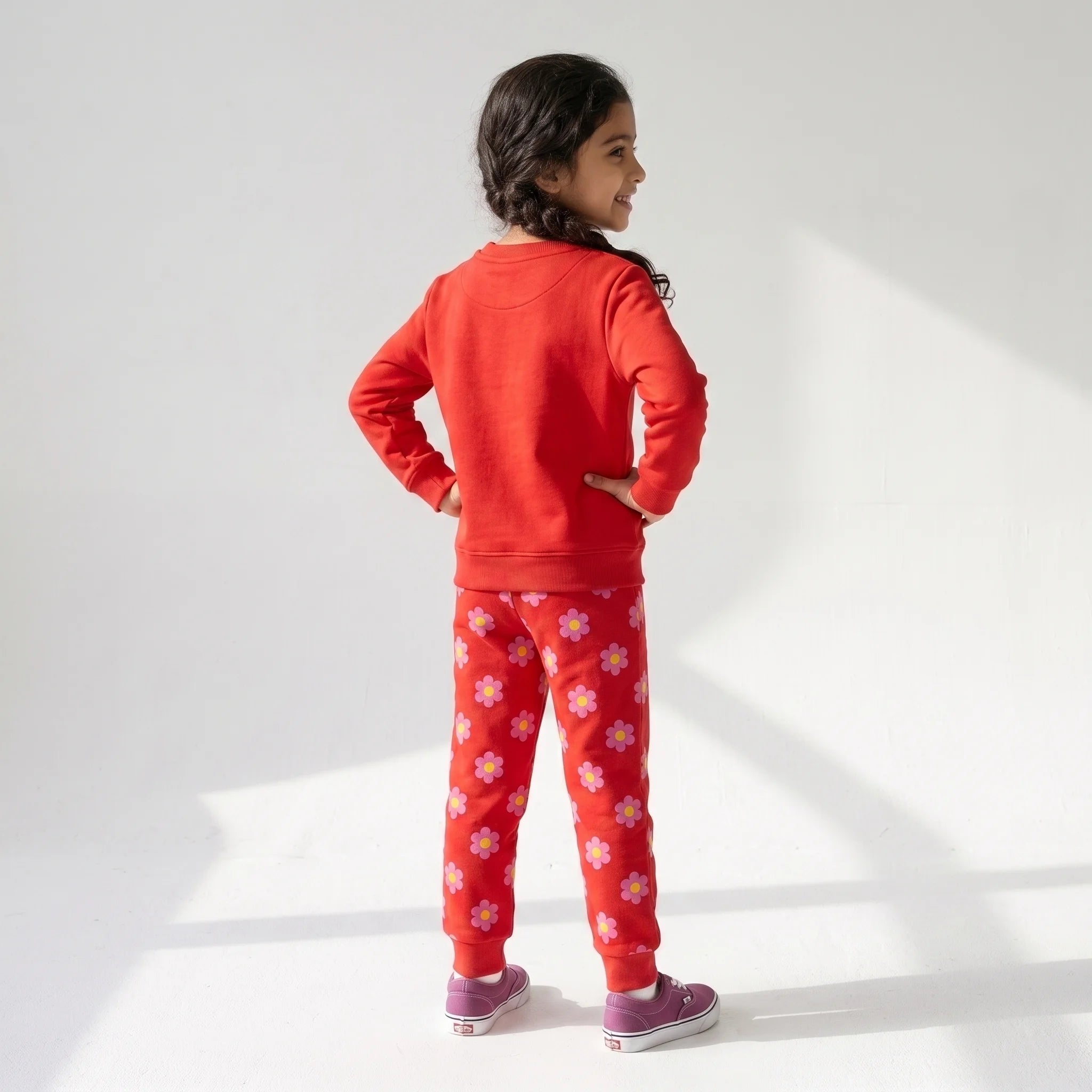 A young girl is shown from behind wearing a red sweatshirt and matching red pants with a white floral pattern, paired with purple sneakers, against a white studio background with soft window-light shadows.