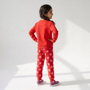 A young girl is shown from behind wearing a red sweatshirt and matching red pants with a white floral pattern, paired with purple sneakers, against a white studio background with soft window-light shadows.