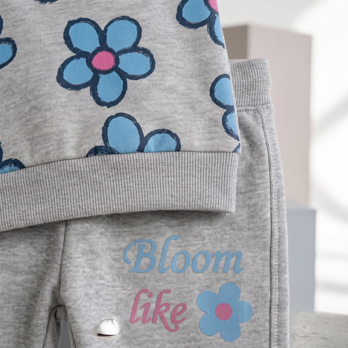 A close-up of a children's gray heathered outfit featuring a top with a blue and pink flower pattern and matching gray pants with the words 'Bloom like' in blue and pink script next to a blue flower graphic.