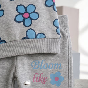 A close-up of a children's gray heathered outfit featuring a top with a blue and pink flower pattern and matching gray pants with the words 'Bloom like' in blue and pink script next to a blue flower graphic.