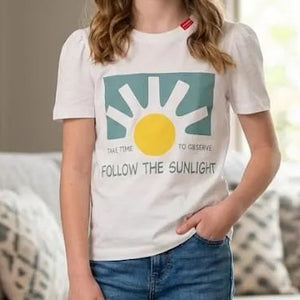 A young girl stands in a bright living room wearing a white graphic t-shirt with a sun design and the text 'FOLLOW THE SUNLIGHT', paired with blue jeans and white sneakers.
