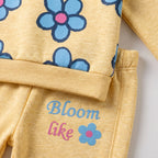 A close-up of a children's two-piece outfit in heathered pale yellow, featuring a long-sleeve top with blue flowers and the pants printed with the words 'Bloom like' in blue and pink alongside a small flower graphic.