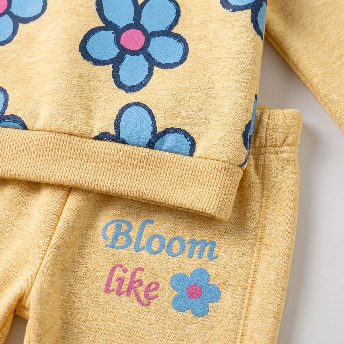 A close-up of a children's two-piece outfit in heathered pale yellow, featuring a long-sleeve top with blue flowers and the pants printed with the words 'Bloom like' in blue and pink alongside a small flower graphic.
