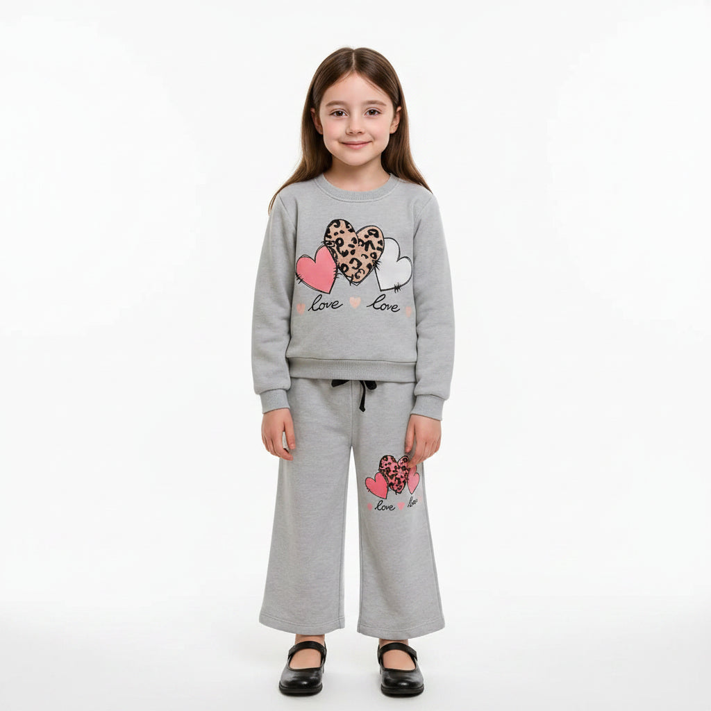A young girl stands indoors wearing a light gray two-piece sweatshirt and jogger pants set with heart and leopard-print graphic details. The background shows a neutral wall and wooden staircase.