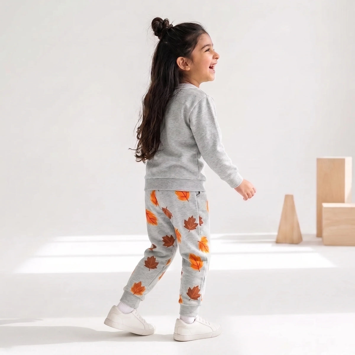 A young girl walking in profile wearing a light gray sweatshirt and matching sweatpants with an orange and brown autumn leaf pattern, paired with white sneakers, in a bright studio setting.