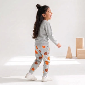 A young girl walking in profile wearing a light gray sweatshirt and matching sweatpants with an orange and brown autumn leaf pattern, paired with white sneakers, in a bright studio setting.