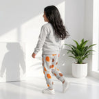 A child wearing a light gray sweatshirt and matching sweatpants with an orange and brown autumn leaf print, walking in a bright white room with a potted plant.