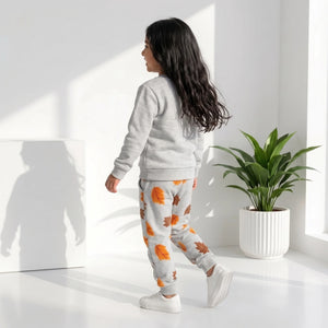 A child wearing a light gray sweatshirt and matching sweatpants with an orange and brown autumn leaf print, walking in a bright white room with a potted plant.