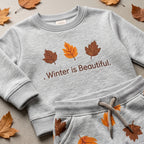 A light heather gray children's sweatshirt and matching sweatpants set laid flat on a textured surface with scattered autumn leaves. The sweatshirt has three printed leaves in brown, orange, and rust tones above the phrase '.