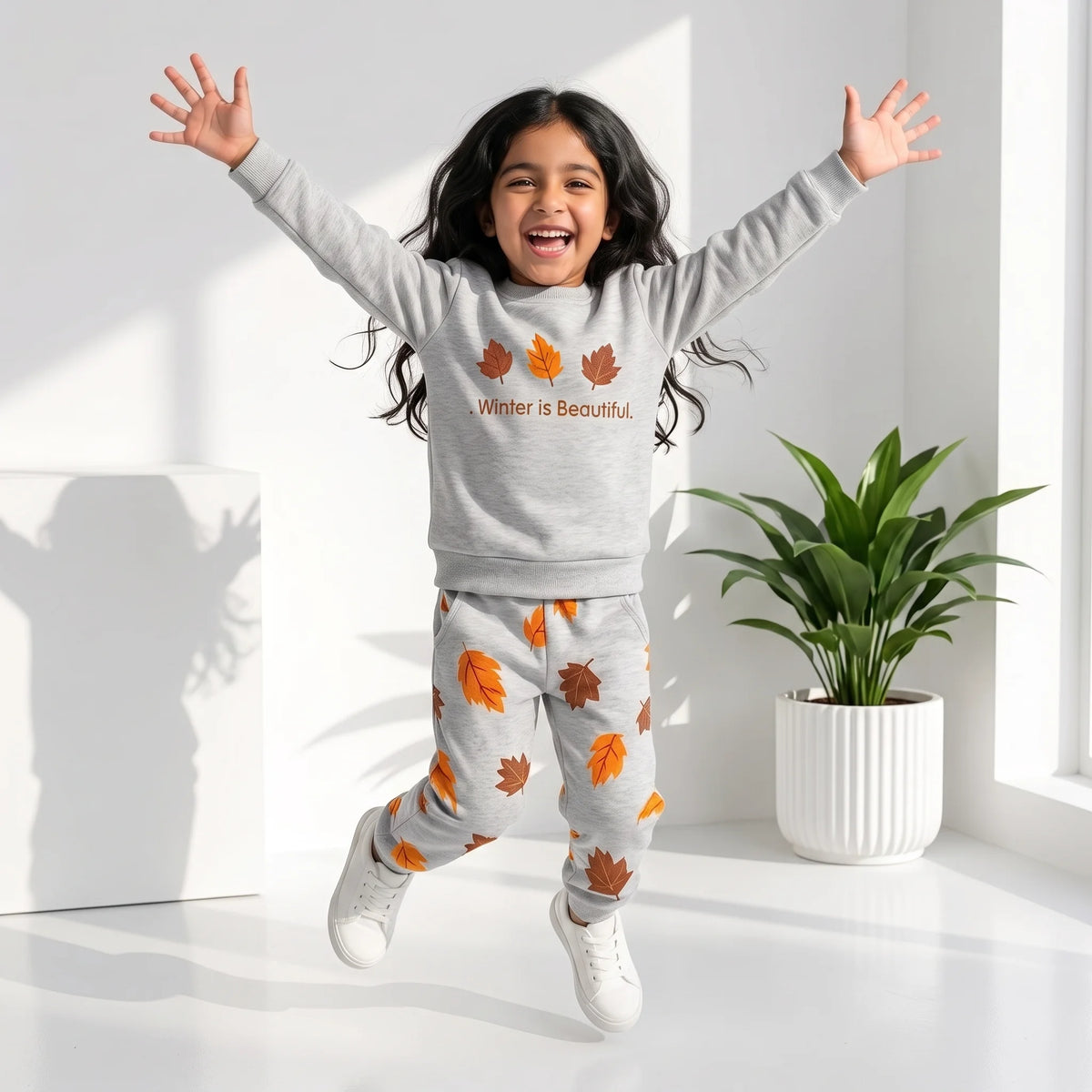 A young girl jumps happily in a bright room wearing a light gray sweatshirt and matching sweatpants with orange and brown autumn leaf prints and the text 'Winter is Beautiful'.