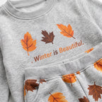 A close-up of a light heather gray sweatshirt with a matching pair of pants. The sweatshirt features a graphic print of three autumn leaves in orange and brown next to the text '.