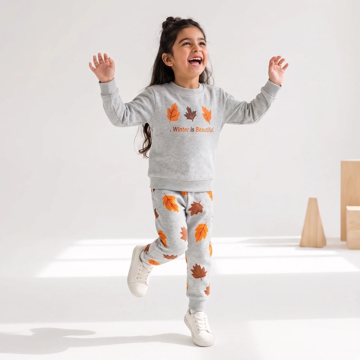 A young girl is captured mid-jump wearing a light gray heathered two-piece pajama set. The sweatshirt features orange and brown autumn leaves and the text 'Winter is Beautiful' in orange.