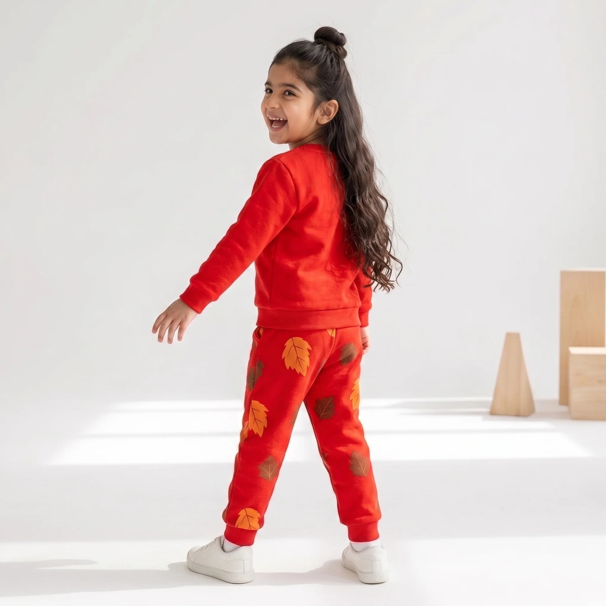 A young girl with long dark hair in a high ponytail is wearing a bright red two-piece sweatshirt and jogger pants set with orange and yellow autumn leaf prints. She is standing in a bright, white studio setting with light wood blocks in the background.