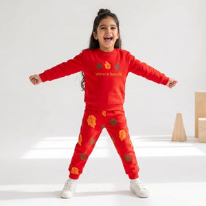 A young girl stands with arms outstretched wearing a bright red two-piece sweatshirt and jogger set with autumn leaf prints and the text 'Winter is Beautiful' on the chest, photographed in a clean white studio setting.