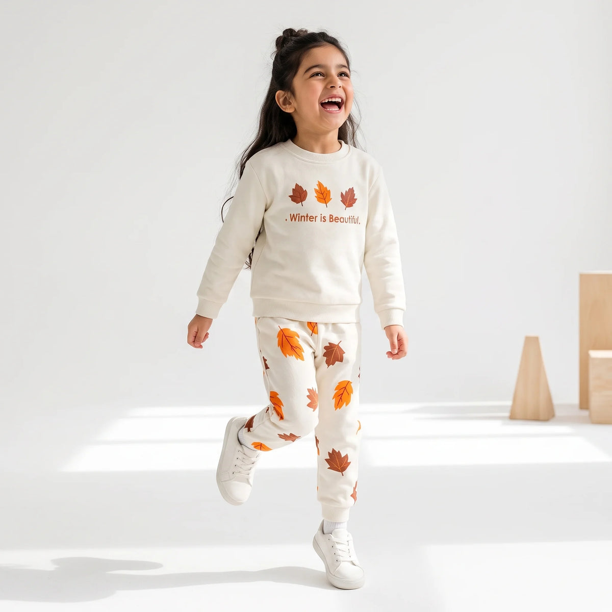 A young girl smiling and running in a bright studio while wearing a cream-colored sweatshirt and matching joggers with orange and brown autumn leaf prints. The sweatshirt features the text 'Winter is Beautiful' under three leaf graphics.