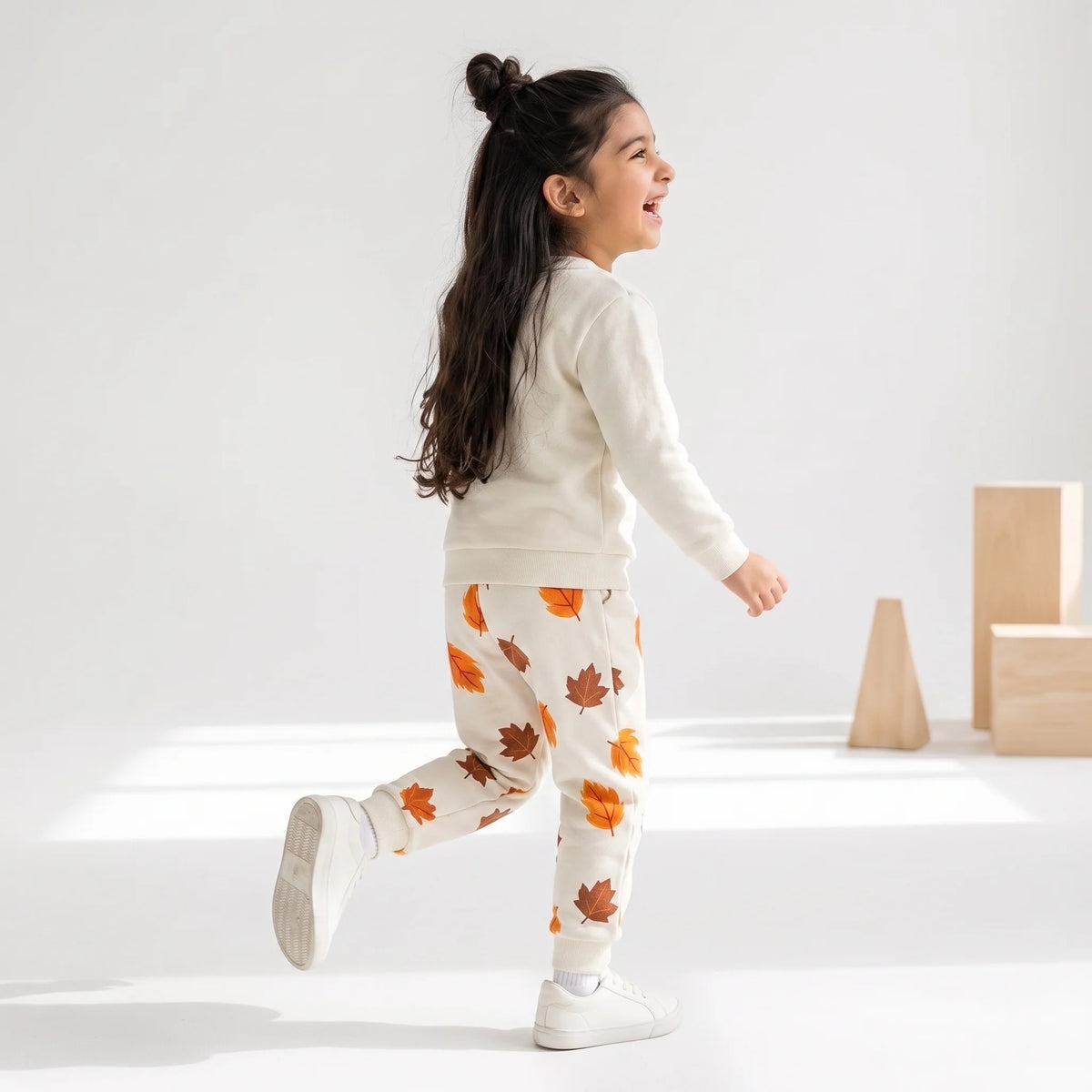 A young girl with her hair in a high bun is shown from the back, wearing a cream-colored sweatshirt and matching sweatpants with an orange and brown autumn leaf pattern. She is walking in a bright, minimalist studio setting with light wood blocks in the background.